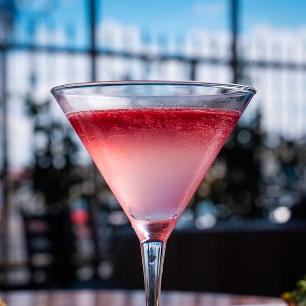 A pink martini with a red top layer and condensation on the glass, set against a blurred window background.