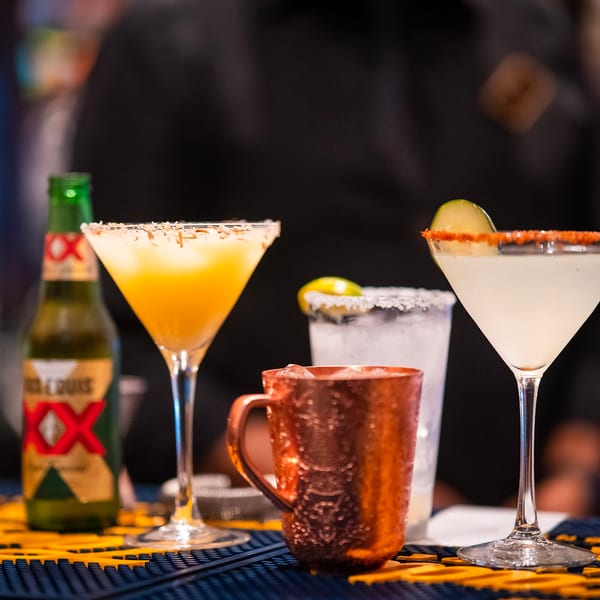 A bar counter with a beer bottle, a Moscow mule, and two margaritas with salt and chili rims.