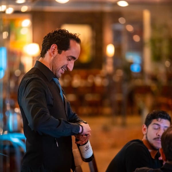 A smiling waiter in a black vest and blue tie holds a wine bottle, serving guests at a restaurant.