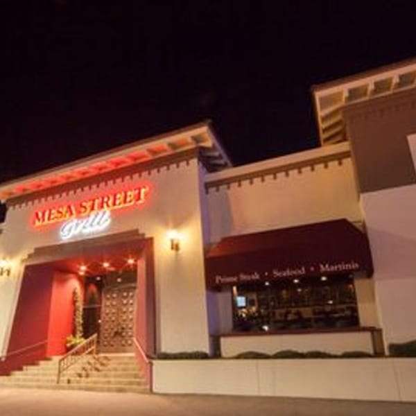 Exterior of Mesa Street Grill at night, with red neon sign and awnings, and illuminated entrance.