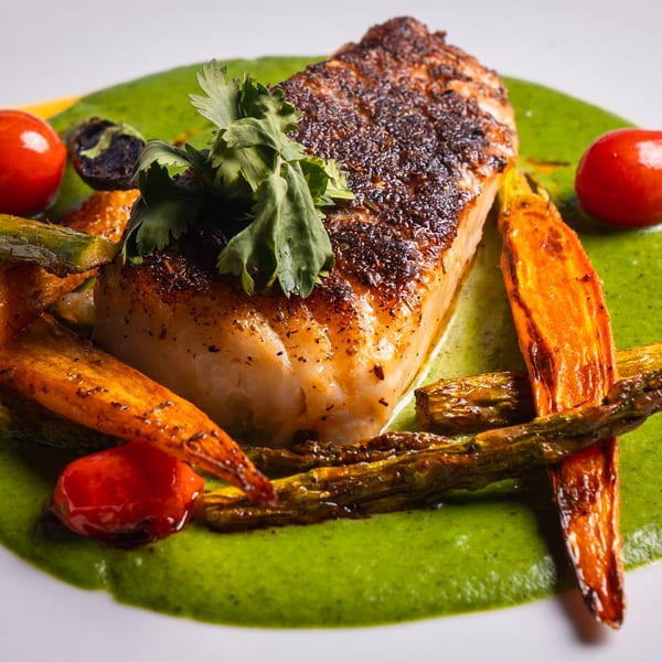 Pan-seared fish with roasted carrots, asparagus, cherry tomatoes, and green sauce on a white plate.