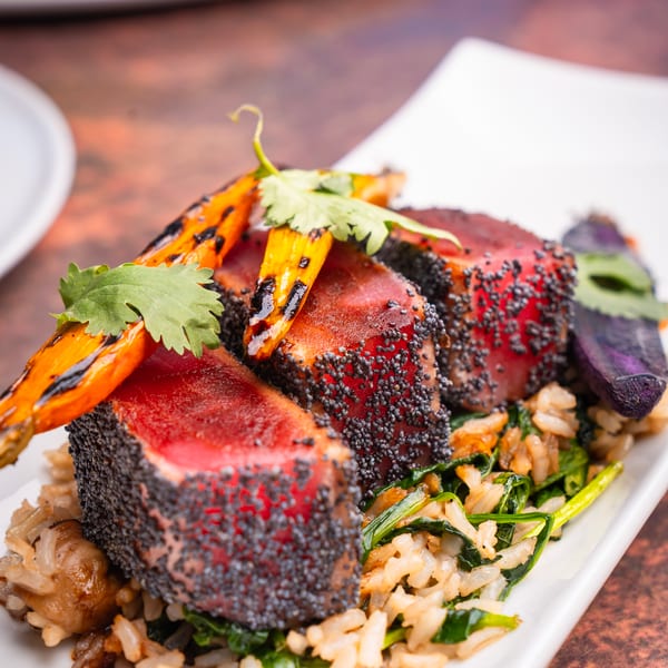 Seared tuna with poppy seeds, grilled carrots, cilantro, and rice with greens on a white plate.