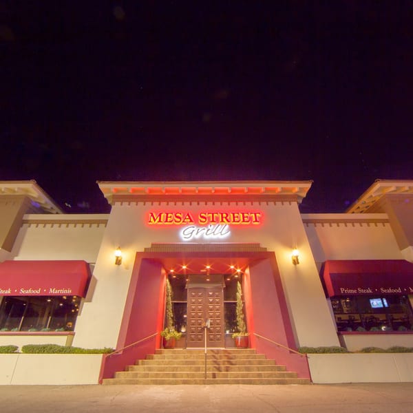 Nighttime exterior of Mesa Street Grill, featuring illuminated signage, red awnings, and a grand staircase entrance.