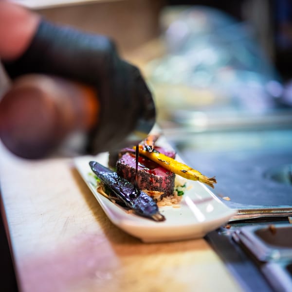 Chef's hand in black glove drizzles sauce over seared tuna with roasted vegetables on a white plate.