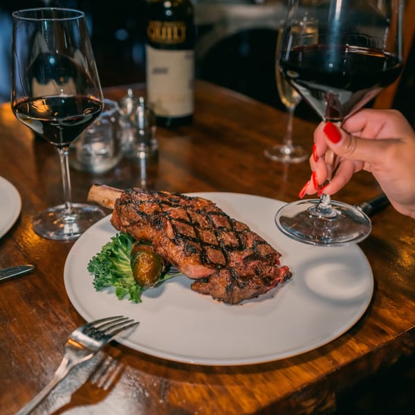 A grilled steak on a white plate with greens, a wine bottle, and a person holding a wine glass.
