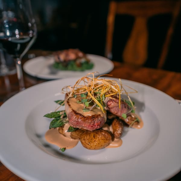 Two plates of steak with asparagus, potatoes, sauce, and crispy garnish on a wooden table.