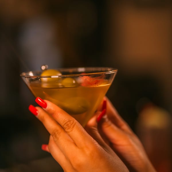 A person with red manicured nails holding a martini glass with olives and a red garnish.