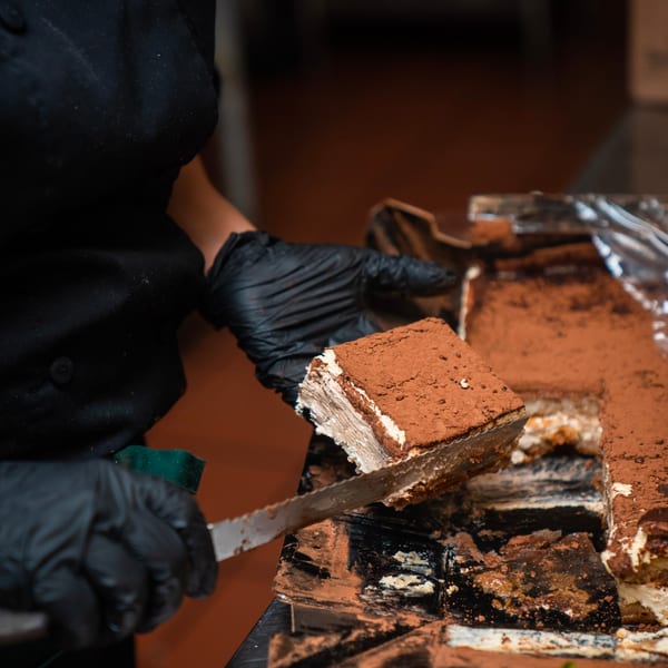 Gloved pastry chef lifts a slice of cocoa-dusted layered chocolate cake from a baking tray.