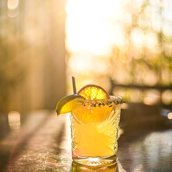 A vibrant yellow cocktail with a lime wedge and orange slice, backlit by golden sunlight.