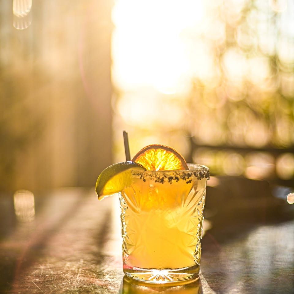 A vibrant yellow cocktail with a lime wedge and orange slice, backlit by golden sunlight.